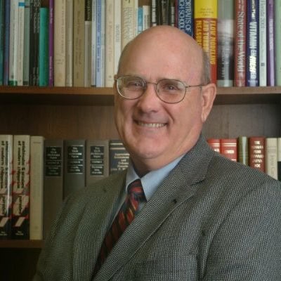 Highly educated man in a professional setting with a bookshelf background, representing academic expertise and institutional knowledge.