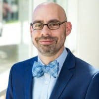 Efficient college student wearing glasses and a blue suit with a bow tie, smiling confidently in a professional setting.