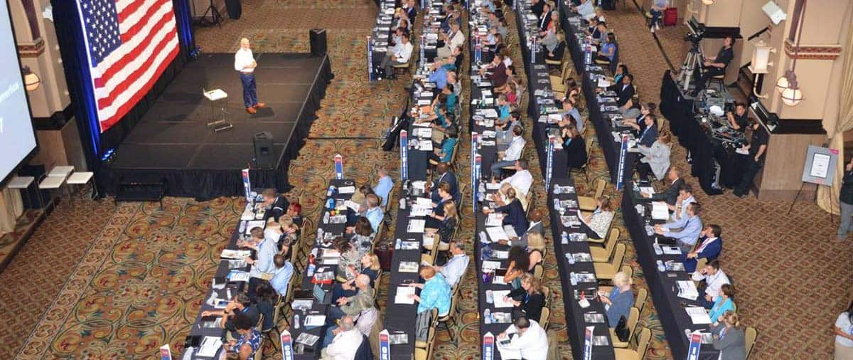 Large conference room filled with attendees seated at long tables, listening to a speaker on stage in front of a large American flag backdrop at a Complete College America event.