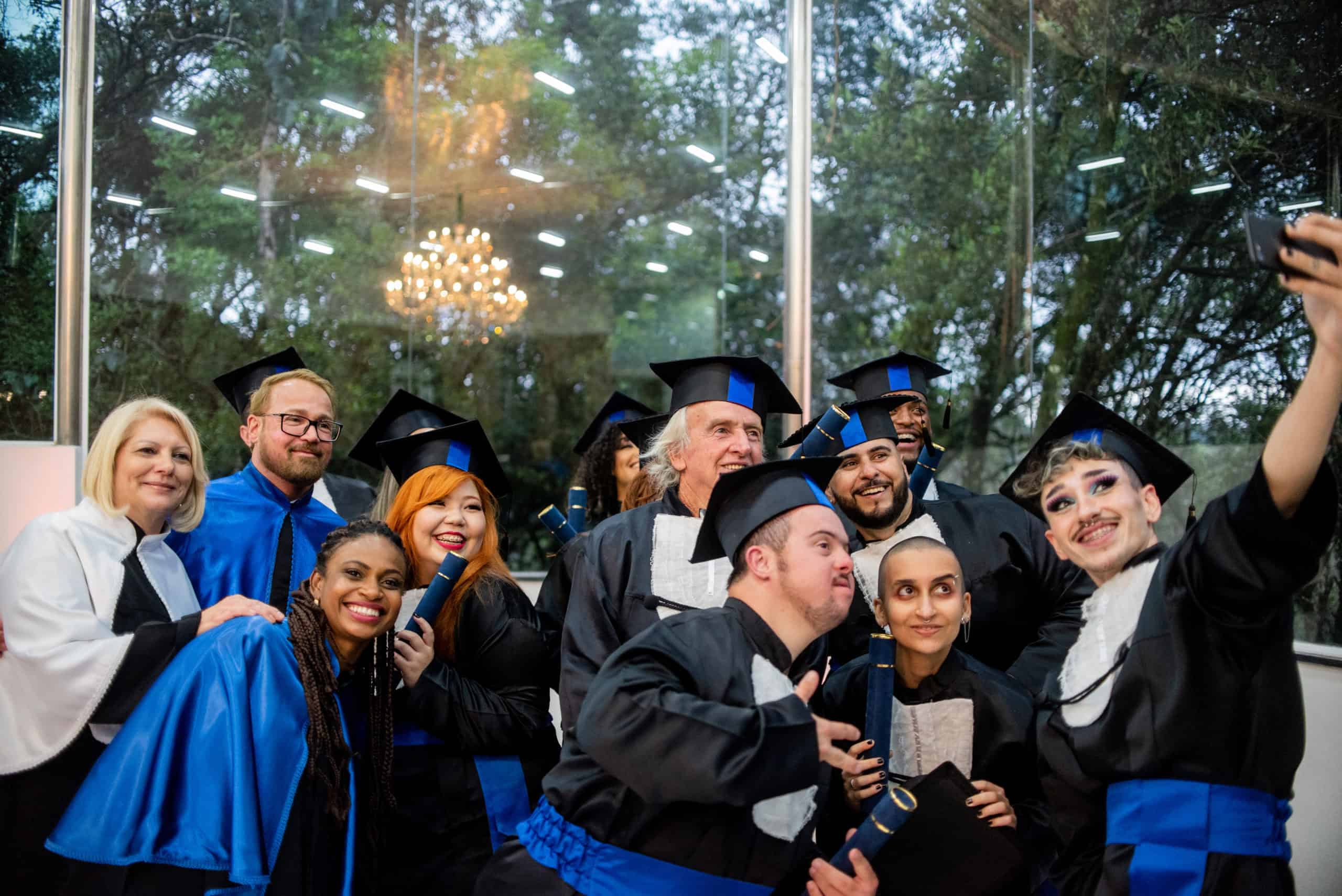 Graduates celebrating at a college graduation ceremony, wearing caps and gowns, with a diverse group taking a selfie inside a modern venue surrounded by trees, emphasizing higher education success.