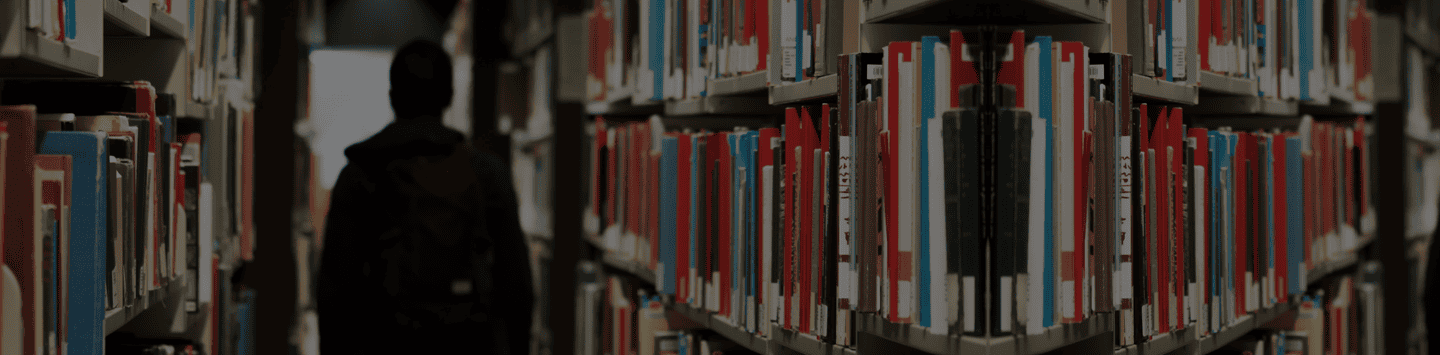 Wide shot of a person browsing books in a library aisle with stacks of colorful books on either side, emphasizing access to higher education and learning resources.