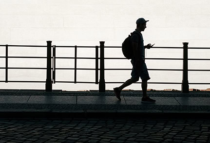 A student walking along a waterfront promenade, using a smartphone, with safety railings in the background.