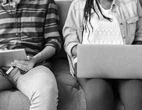 ALT: Two students using laptops and tablets on a sofa, representing college engagement and digital learning.
