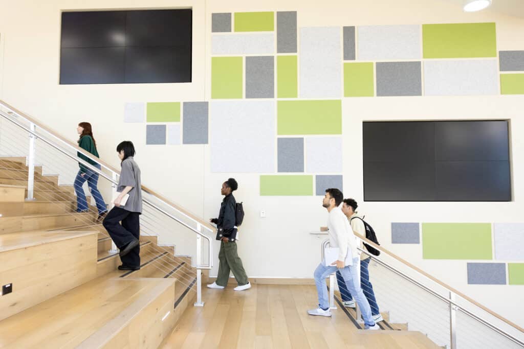 Modern college students ascending wooden stairs inside a contemporary campus building with colorful wall art, emphasizing higher education, student life, and learning environments.