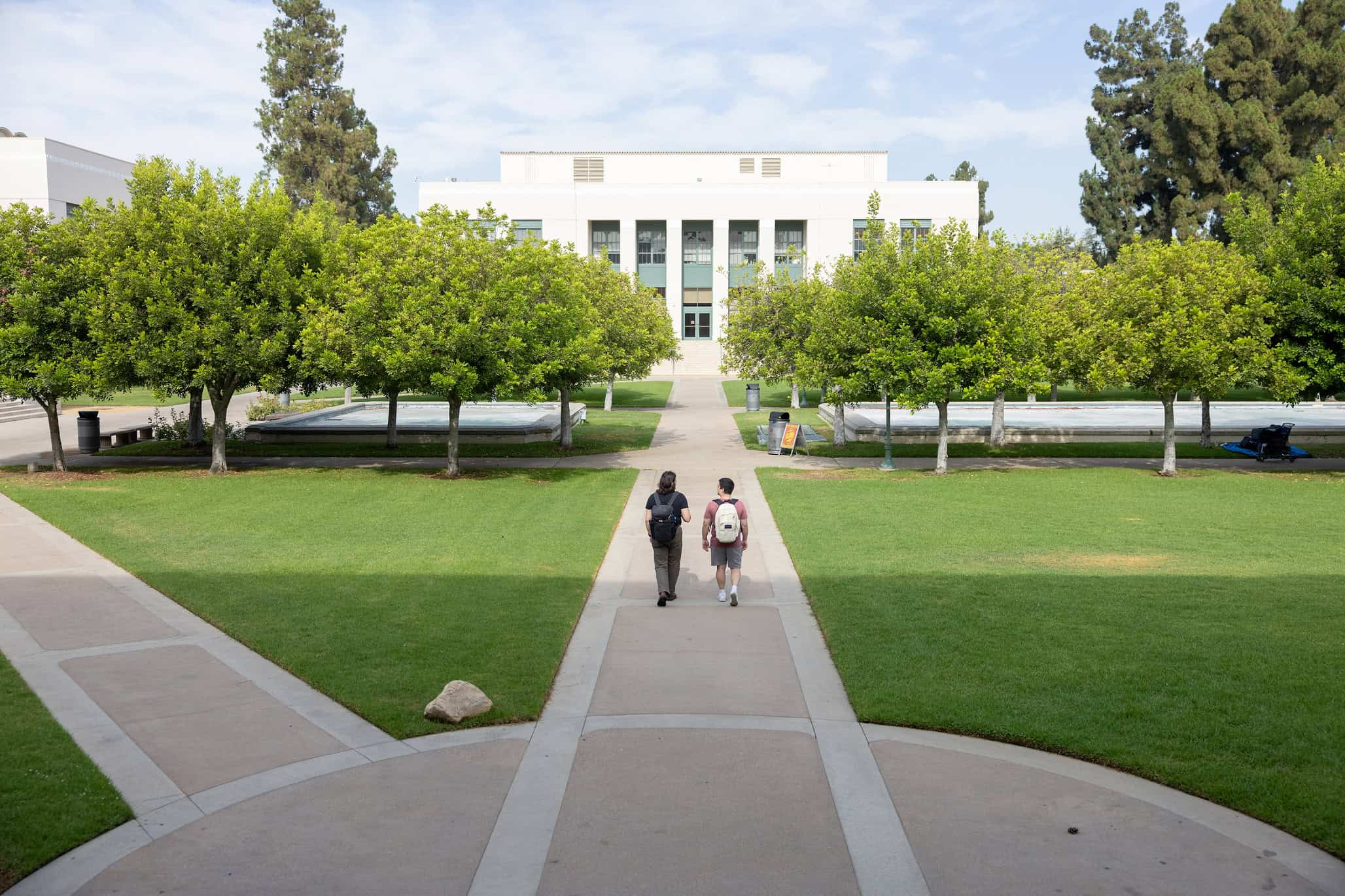 Students walking on campus toward a university building with lush greenery and trees in the background, emphasizing higher education and college environment.