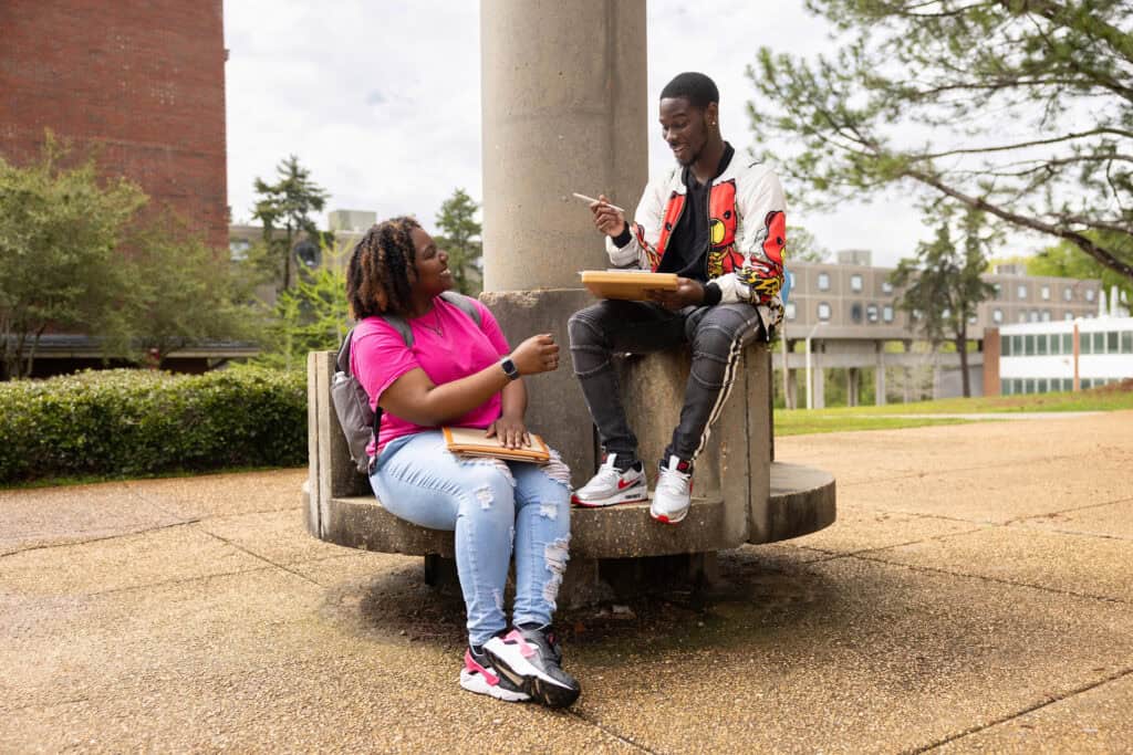 Open college campus scene with two diverse students engaging in a friendly conversation outdoors.