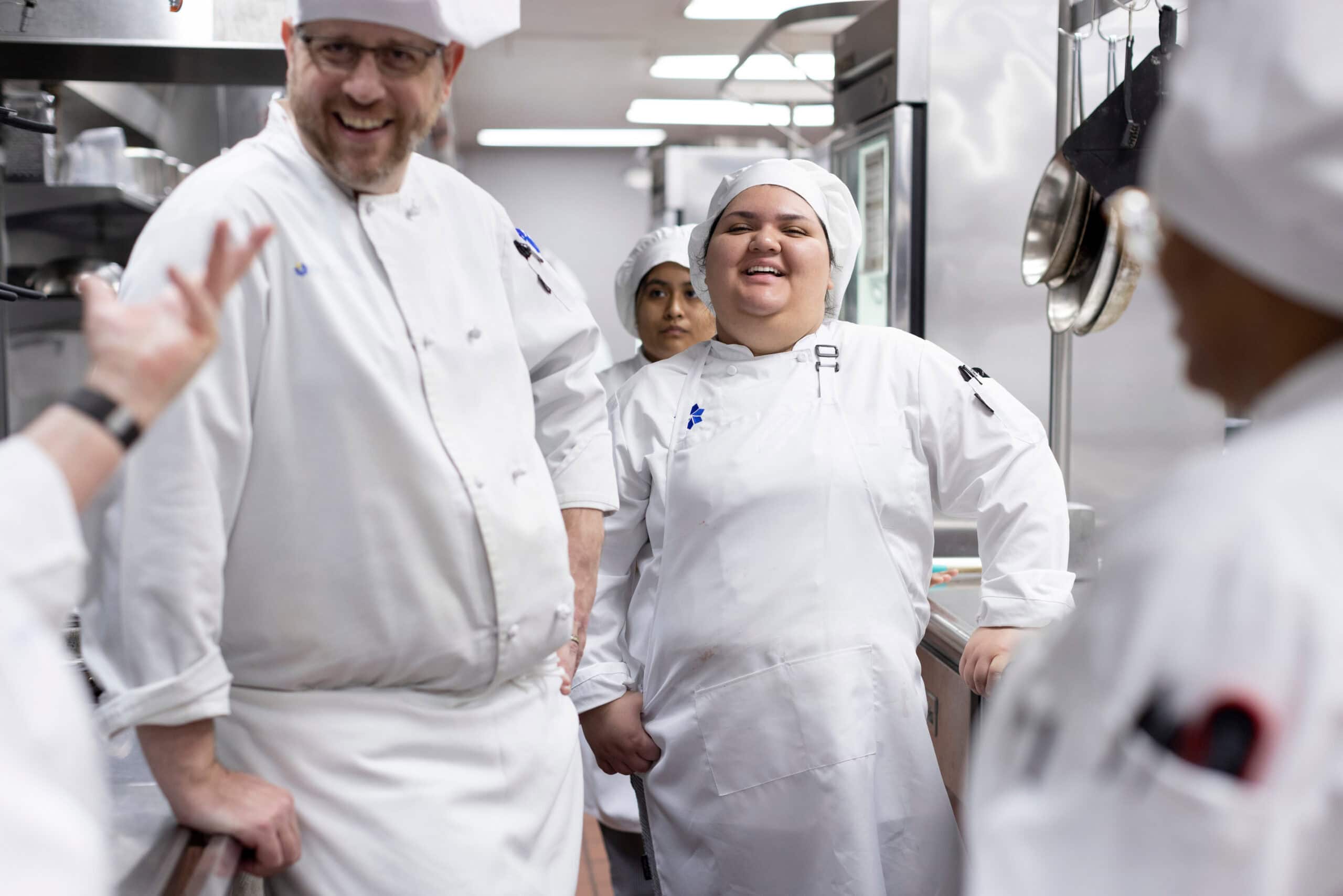 Laughter and teamwork among culinary students and instructors in a professional kitchen setting.