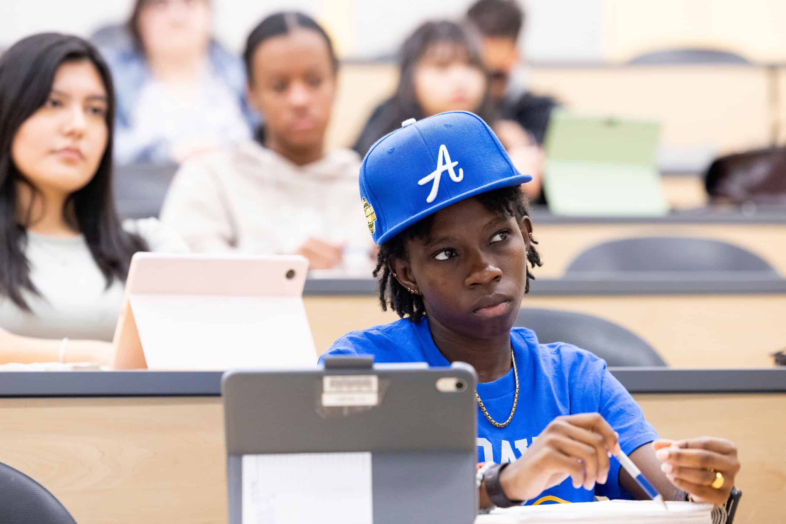 Blue hat with "A" varsity cap, diverse college classroom with attentive students focusing on completing college education, inclusive higher education environment, college success strategies, Complete College America initiative.