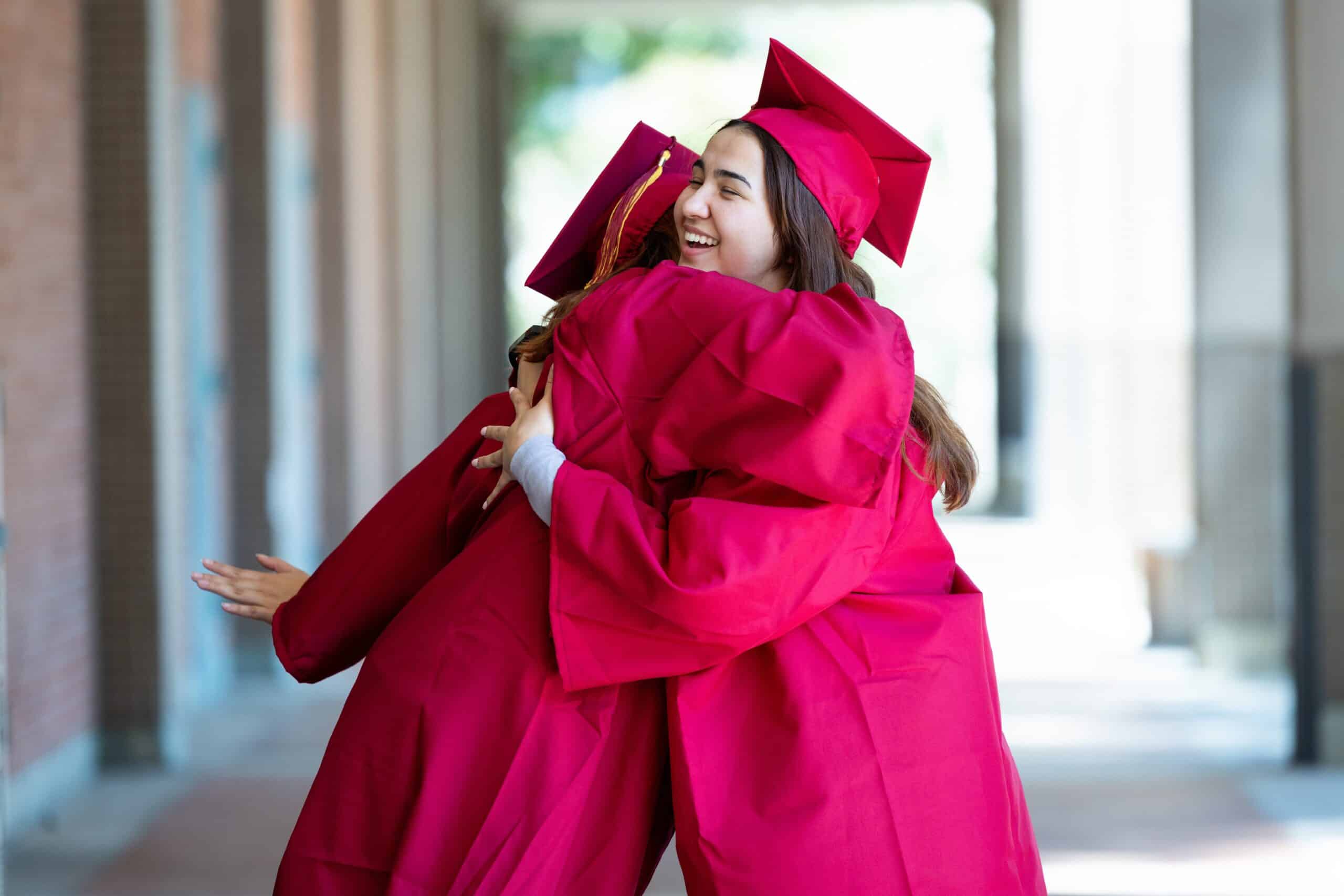 Hugging graduates wearing bright pink caps and gowns outside on a sunny campus corridor.