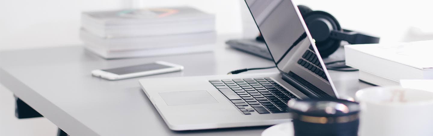 A modern workspace with a silver laptop, smartphone, headphones, and books on a white desk, illustrating themes of higher education, college success, and academic achievement.