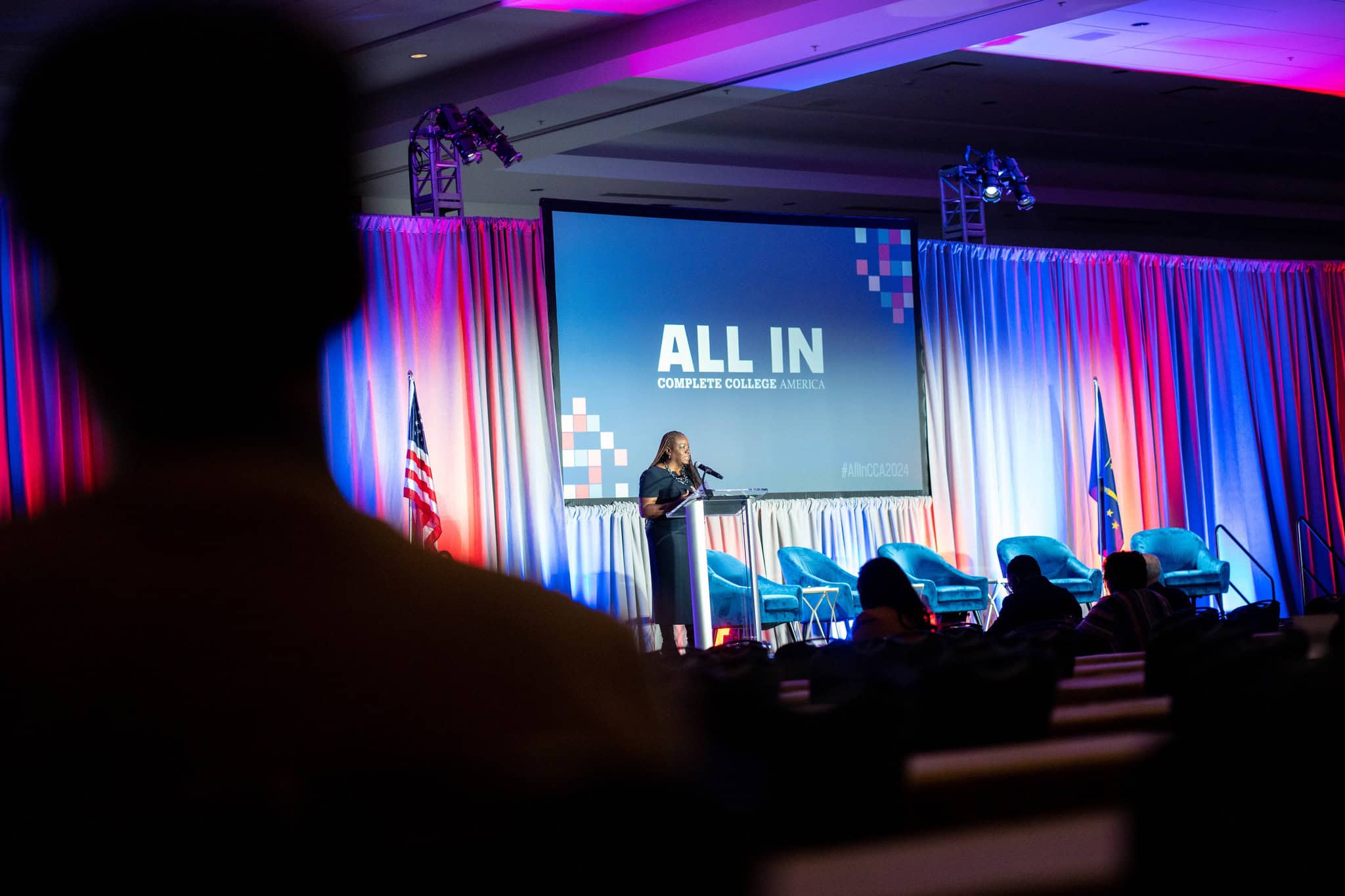 Diverse speaker at a Complete College America event on a stage with a large screen, American flags, and vibrant lighting, emphasizing commitment to increasing college completion and student success.