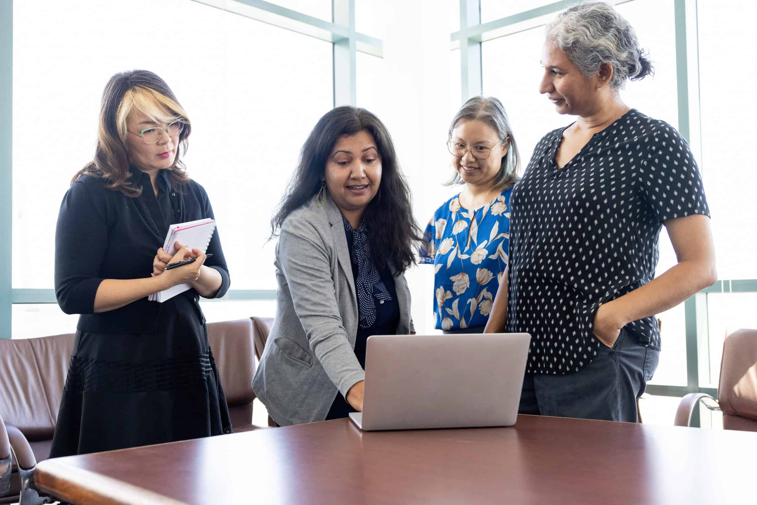 Diverse group of women collaborating around a laptop in a modern office, promoting initiatives for increasing college completion rates and student success.