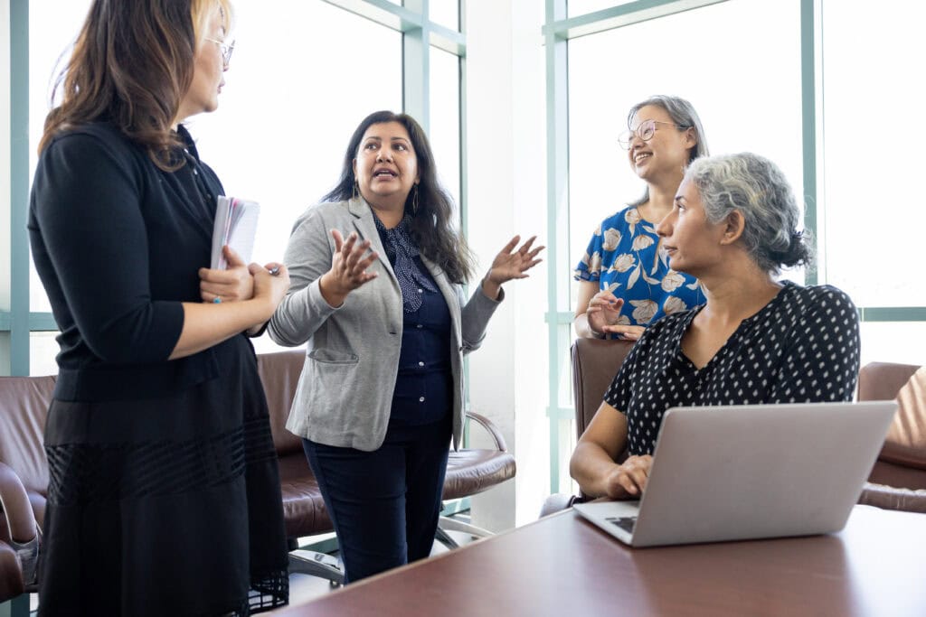 Four Asian female faculty holding a meeting around a laptop computer in a board room on a college campus.